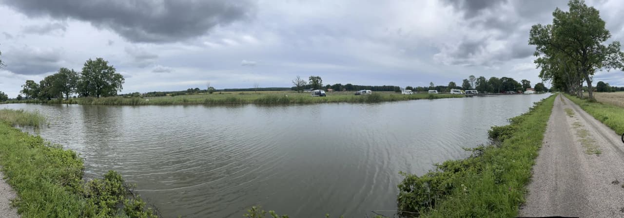 En naturskön vy över Jonsboda med en vattenresurs i förgrunden, omgiven av grönska och träd. Himlen är klart blå med några spridda moln, vilket bidrar till den lugna och fridfulla atmosfären.