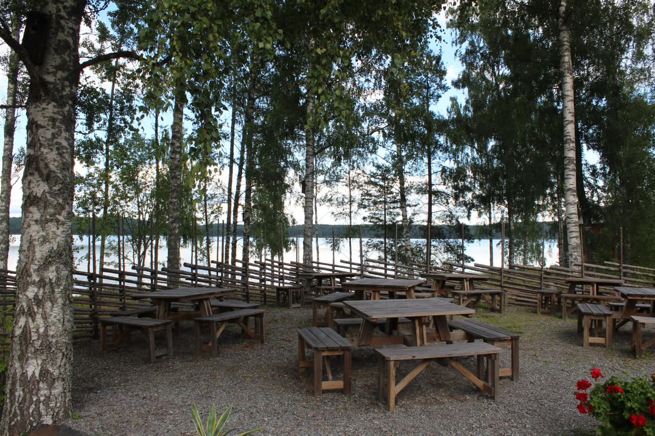 En bild av Älgå hembygdsgård, med ett picknickbord och bänkar i skuggan utomhus, omgiven av grönskande natur och sjöutsikt.