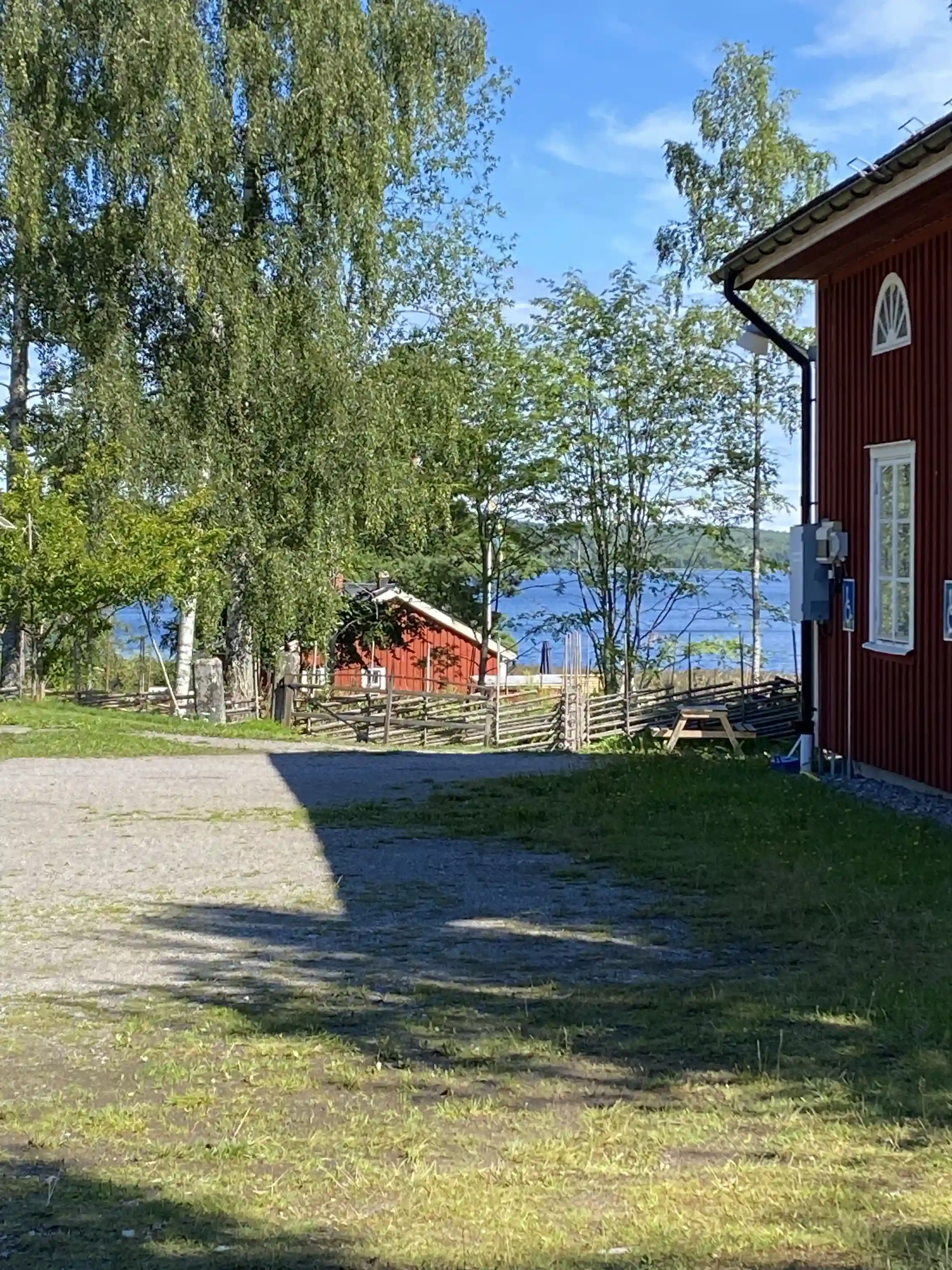 En lantlig stuga omgiven av en grön gräsmatta och träd i skugga på Älgå hembygdsgård. I bakgrunden syns trädgårdsbyggnader och ett parklandskap.