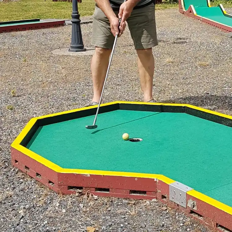 En närbild av en minigolfbana med konstgräs på Gudingebadet, där en person deltar i ett spel. Banan är omgiven av grönska och aktivitetsutrustning, vilket bidrar till en rekreativ och lekfull atmosfär.