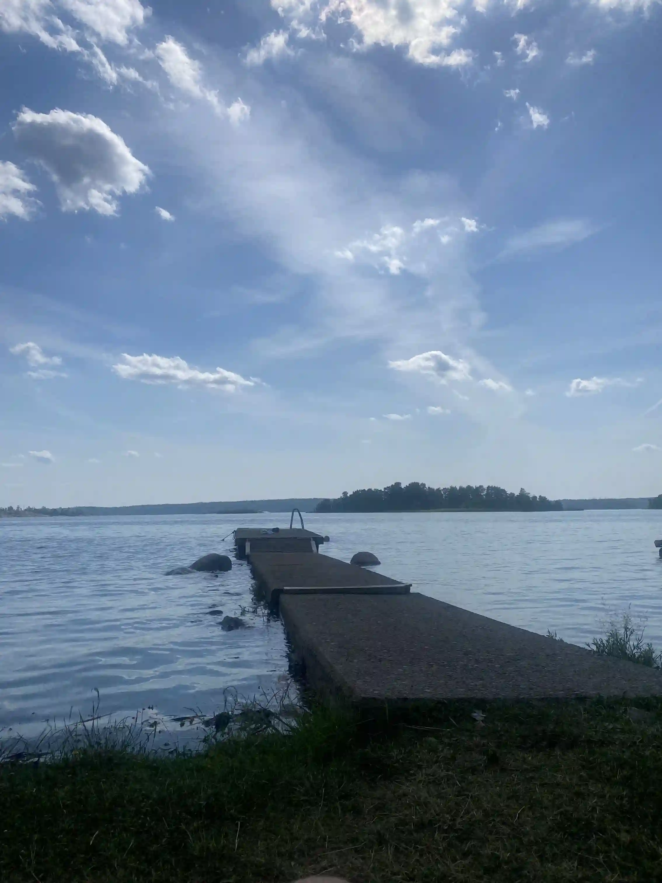 En solig dag vid Gudingebadet med klarblå himmel och vita moln. Ett stort vattenområde sträcker sig mot horisonten, med gröna strandbankar i förgrunden.