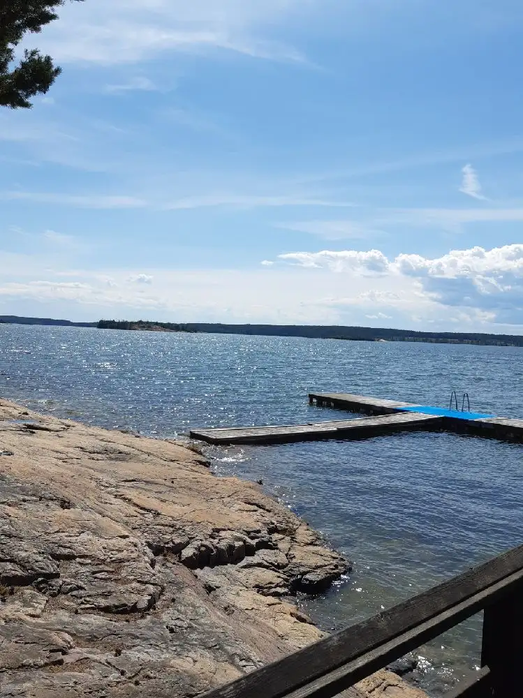 En vidsträckt kustlinje med klart vatten sträcker sig längs Gudingebadet. En badbrygga når ut över det spegelblanka havet och en sandstrand syns vid strandkanten.