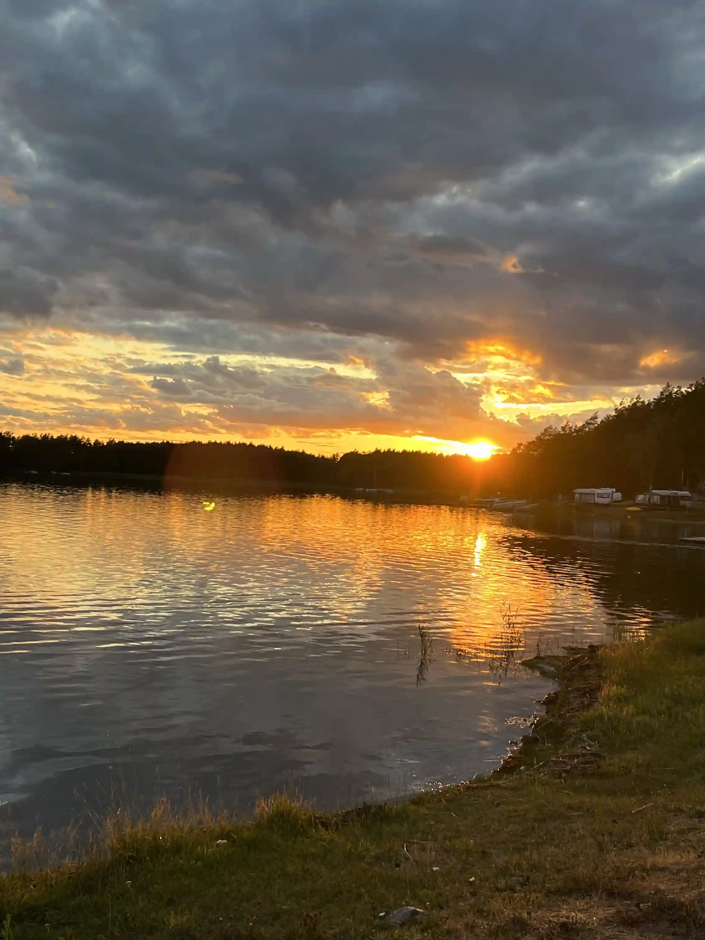 En stilla sjö vid solnedgången, med ett färggrant sken från himlen reflekterande i vattnet. Enstaka moln svävar över horisonten, vilket bidrar till en lugn och fridfull atmosfär vid Gudingebadet.