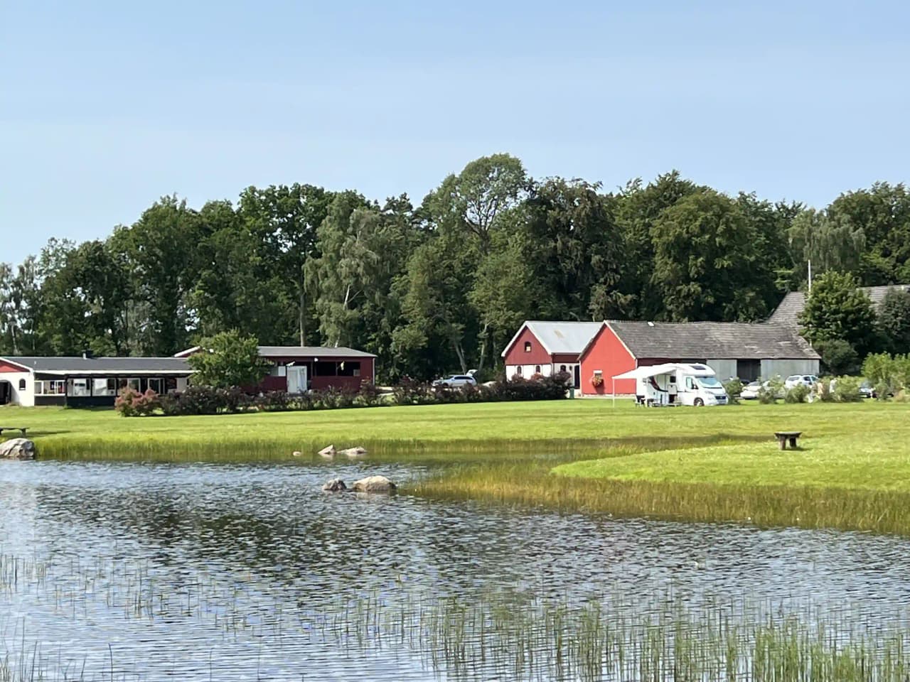 En grön landskapsvy över Vinslövs camping, med vidsträckta gräsmarker och en stilla vattenström. I bakgrunden syns enkla stugor omgivna av lummiga träd, vilket ger en lantlig och fridfull känsla.