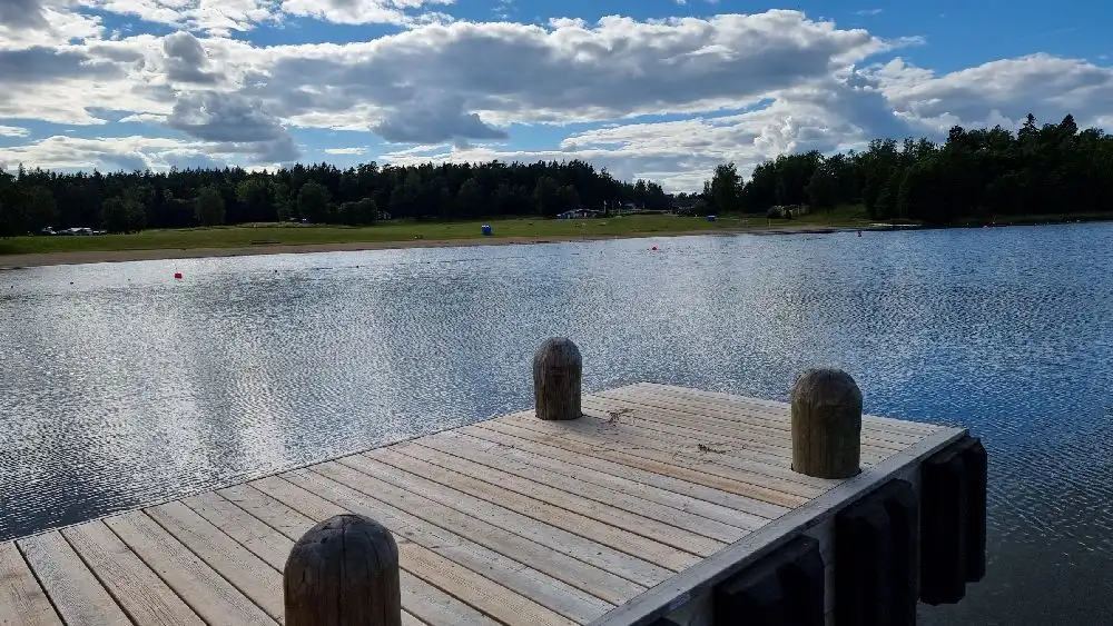 En vy över Farstanäs camping & havsbad visar en lugn vattenyta med molnig himmel i bakgrunden. Vattnet omges av gröna bankar, medan en liten brygga sträcker sig ut i sjön.