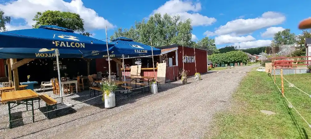 Bild av Gunnagård, som visar ett utomhusområde med picknickbord och paraplyer. I bakgrunden finns parkeringsplats och vägkrogsmiljö, vilket skapar en välkomnande atmosfär för besökare att vila och njuta av en måltid vid de solskyddade borden.