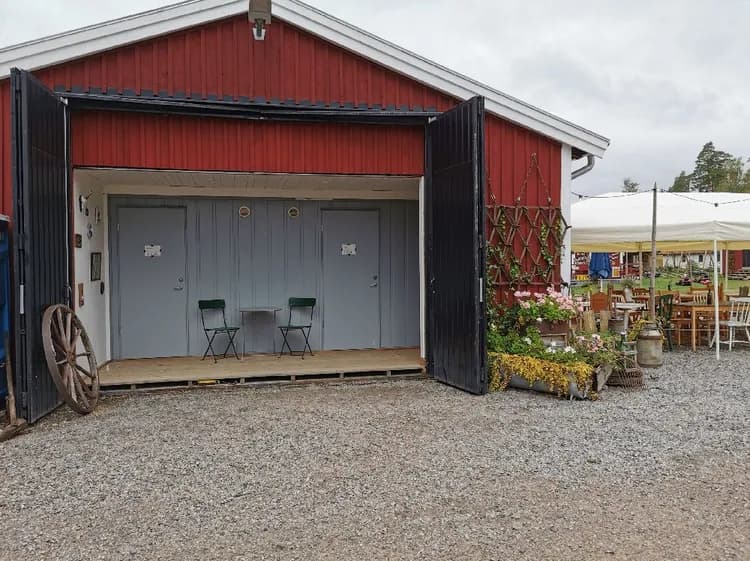 Alt-text: En lantlig gårdsmiljö på Gunnagård med en lada, blomkrukor och andra trädgårdsbyggnader, belägen vid en grusväg.