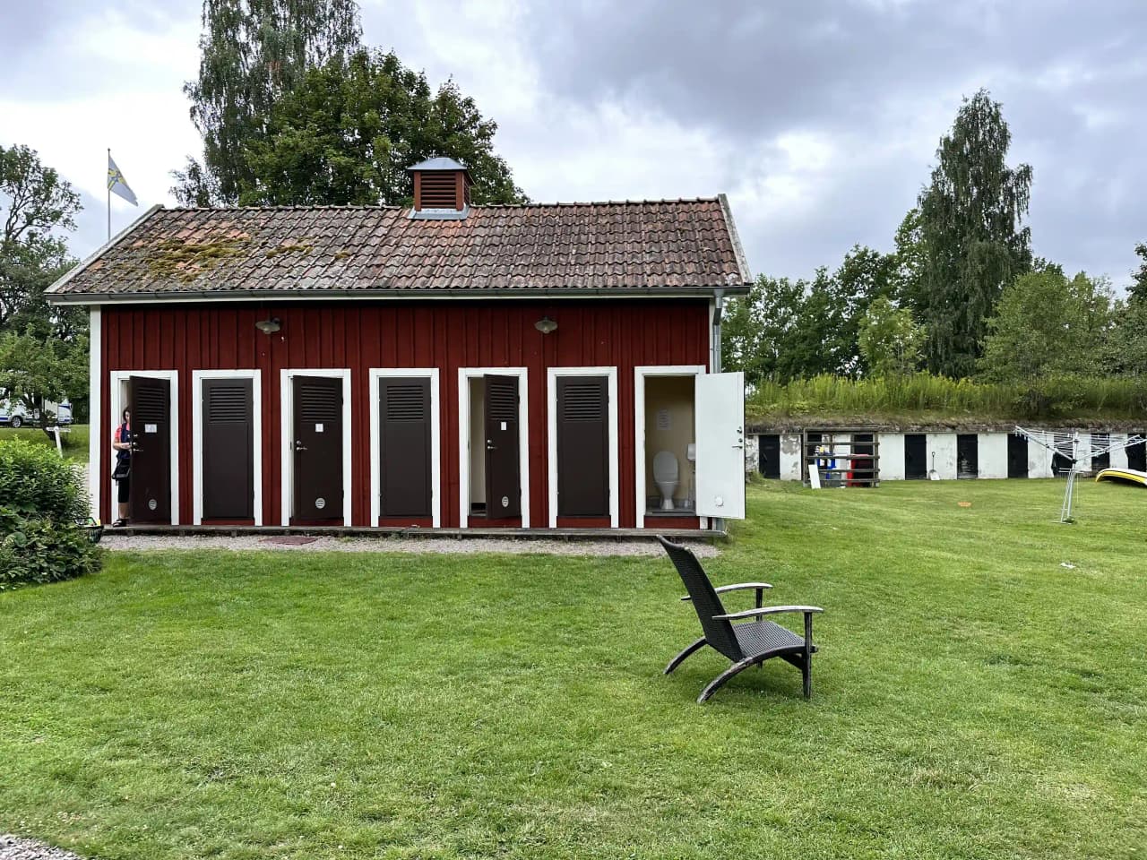 En lummig trädgård med välskött gräsmatta och skuggade områden, omgiven av trädgårdsbyggnader och ett lantligt hus i bakgrunden, vid glasbruket.