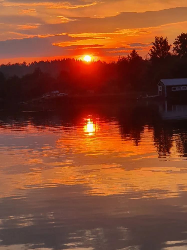 En kvällshimmel över Trångsvikens bygdegård, med orange solnedgång och moln som reflekteras på vattnet i förgrunden.