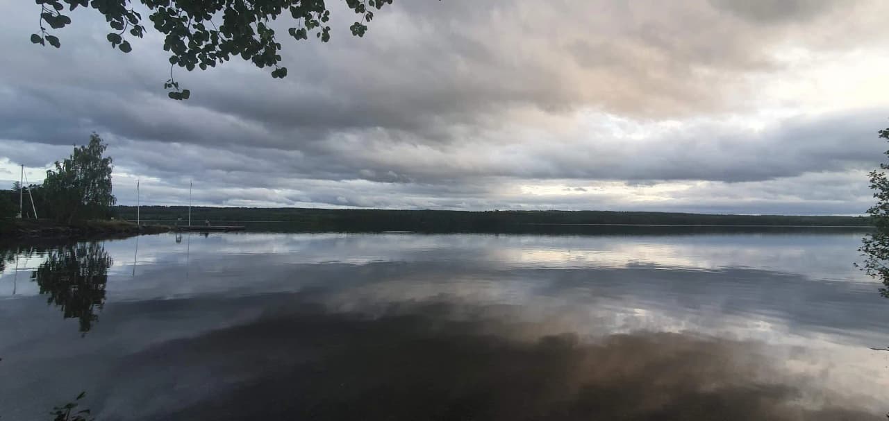 En blå himmel med moln speglas i en lugn vattensamling, omgiven av natur och en vidsträckt horisont.
