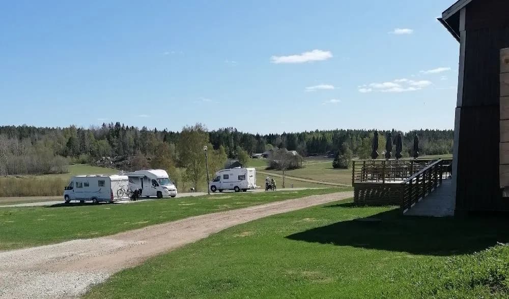 En rymlig äng med husvagnar parkerade bredvid varandra, omgiven av grönskande ängar och tallar, på en plats som erbjuder avkoppling och naturupplevelser.