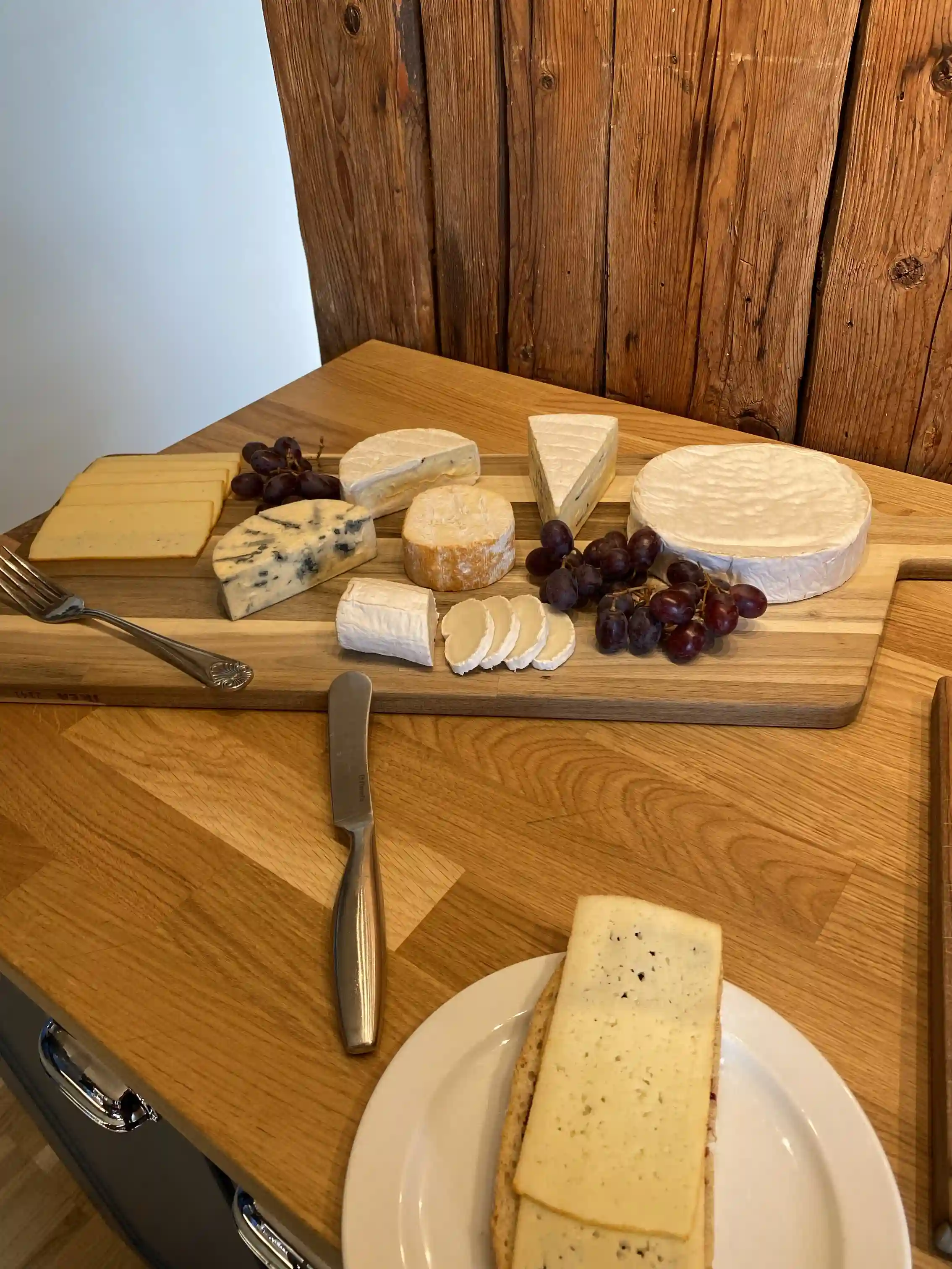 En träskärbräda med olika sorters ost, frukt och köksredskap, placerad på ett dukat bord.