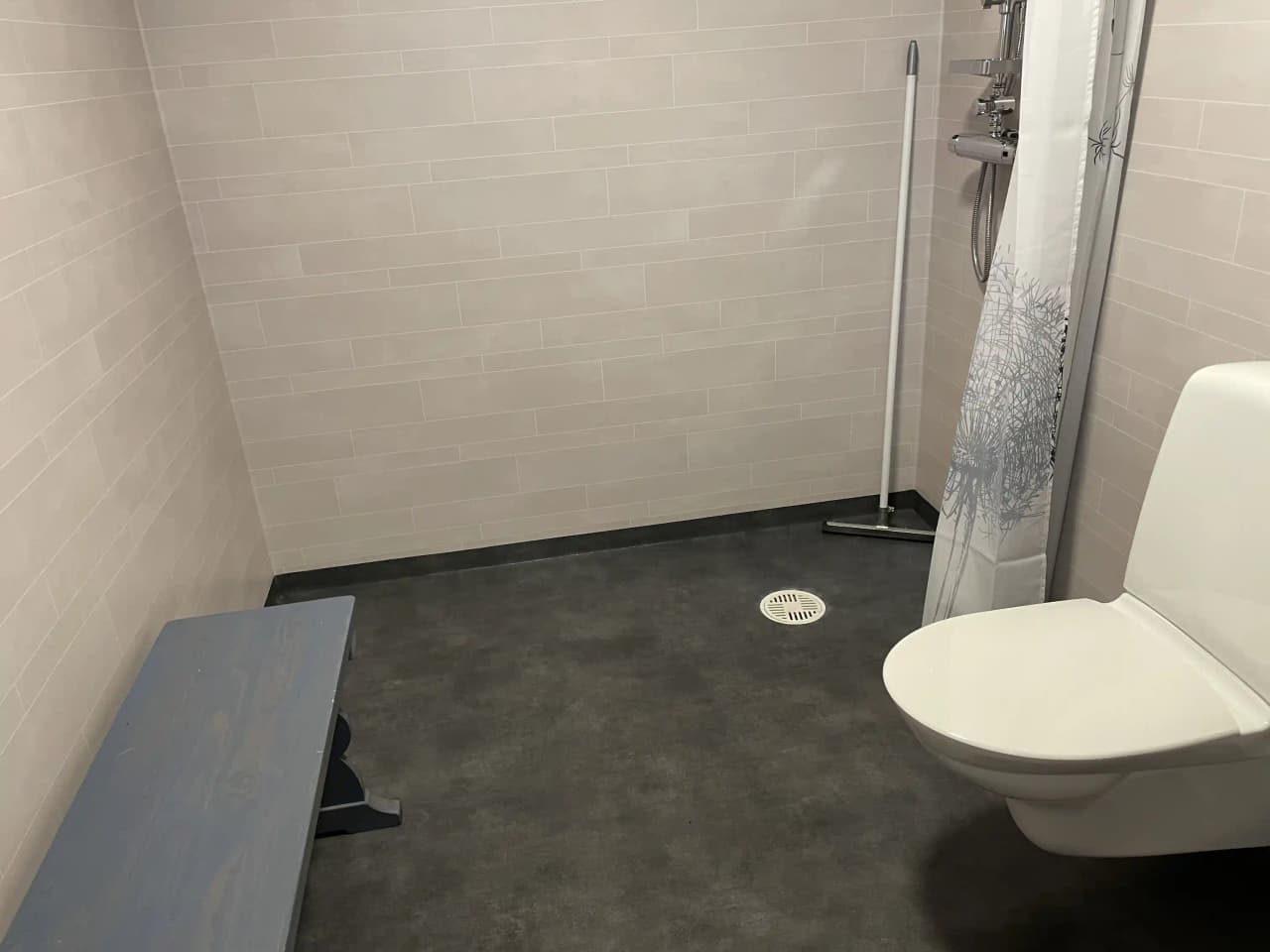 En ljus och modern badrumsmiljö med grått kakelgolv. I fokus står en vit toalett med säte mot en vägg. Kakelplattorna fortsätter upp på väggen och ger ett stilrent intryck.