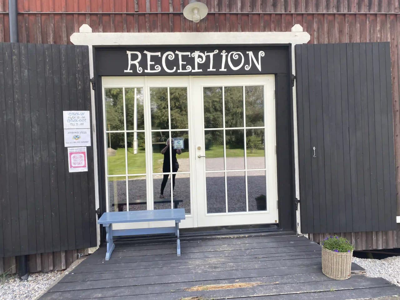 En trästuga med en dörr av mörkbetsat trä. Stugans fasad består av liggande, omålad panel, omgiven av en grönskande gård.