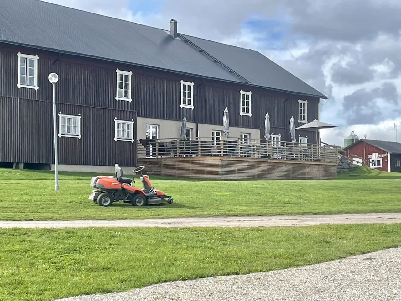 En grön gräsmatta med en liten stuga med träfasad och sadeltak i bakgrunden. Moln syns på himlen över det öppna landskapet. En stolpe står vid sidan om stugan.