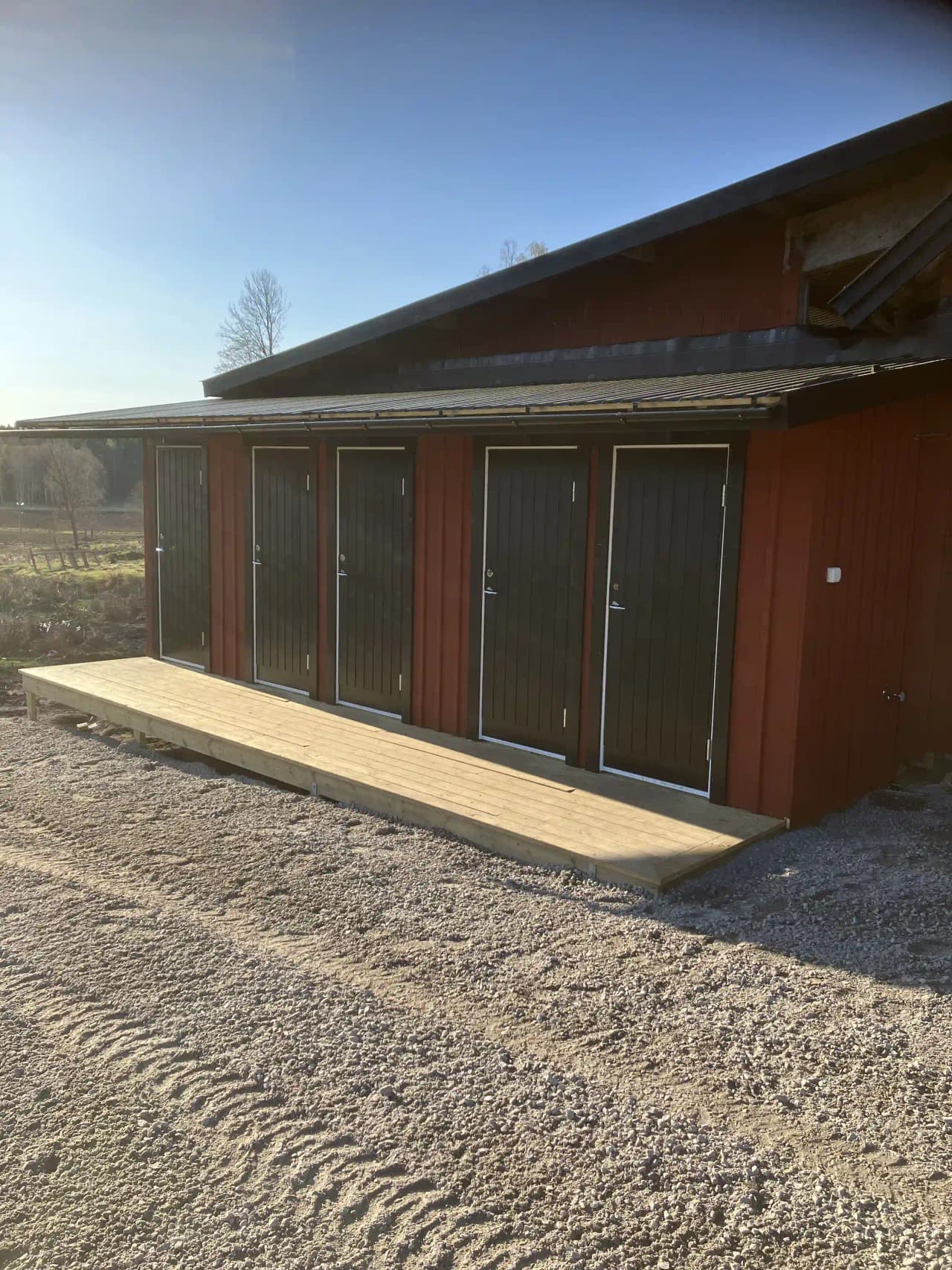 En trästuga vid Billingens camping och stugby, omgiven av grönska, med träpaneler, brunt tak och en enkel dörr. I förgrunden finns en välskött trädgård.