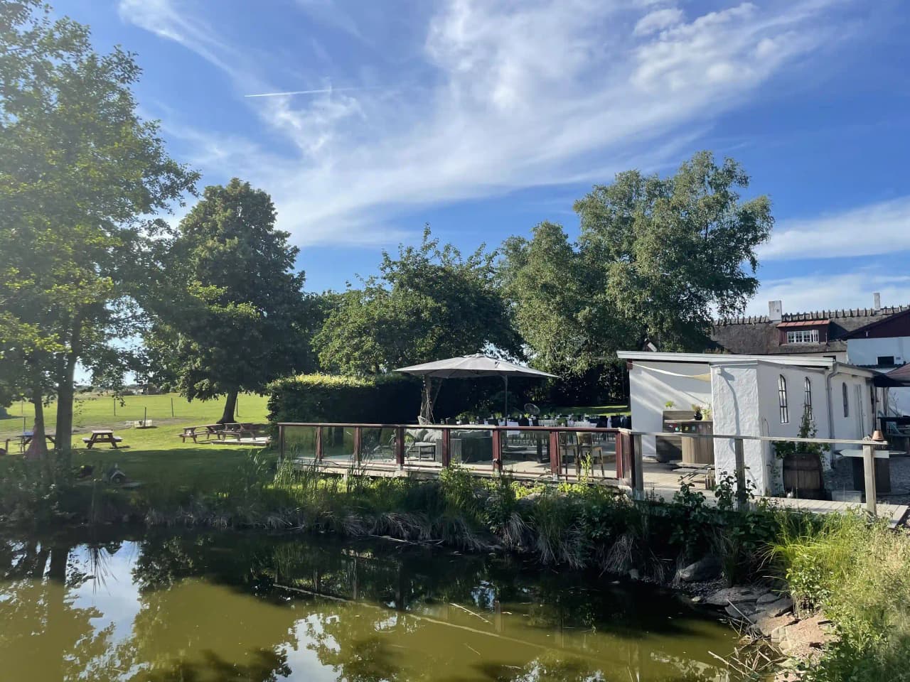 En naturskön vy över Husargården med en damm omgiven av träd och växter. Utemöbler och parasoll är placerade vid vattnet, där en kanal speglas i dammen.