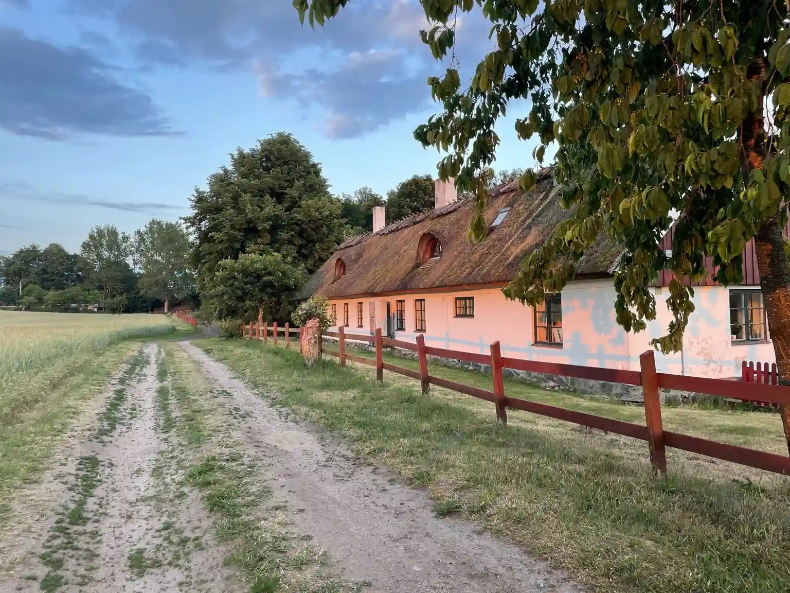 En pittoresk röd stuga omgiven av grönska, belägen på landsbygden vid Husargården. Staket och öppna fält syns i bakgrunden, vilket skapar en lantlig och idyllisk atmosfär.