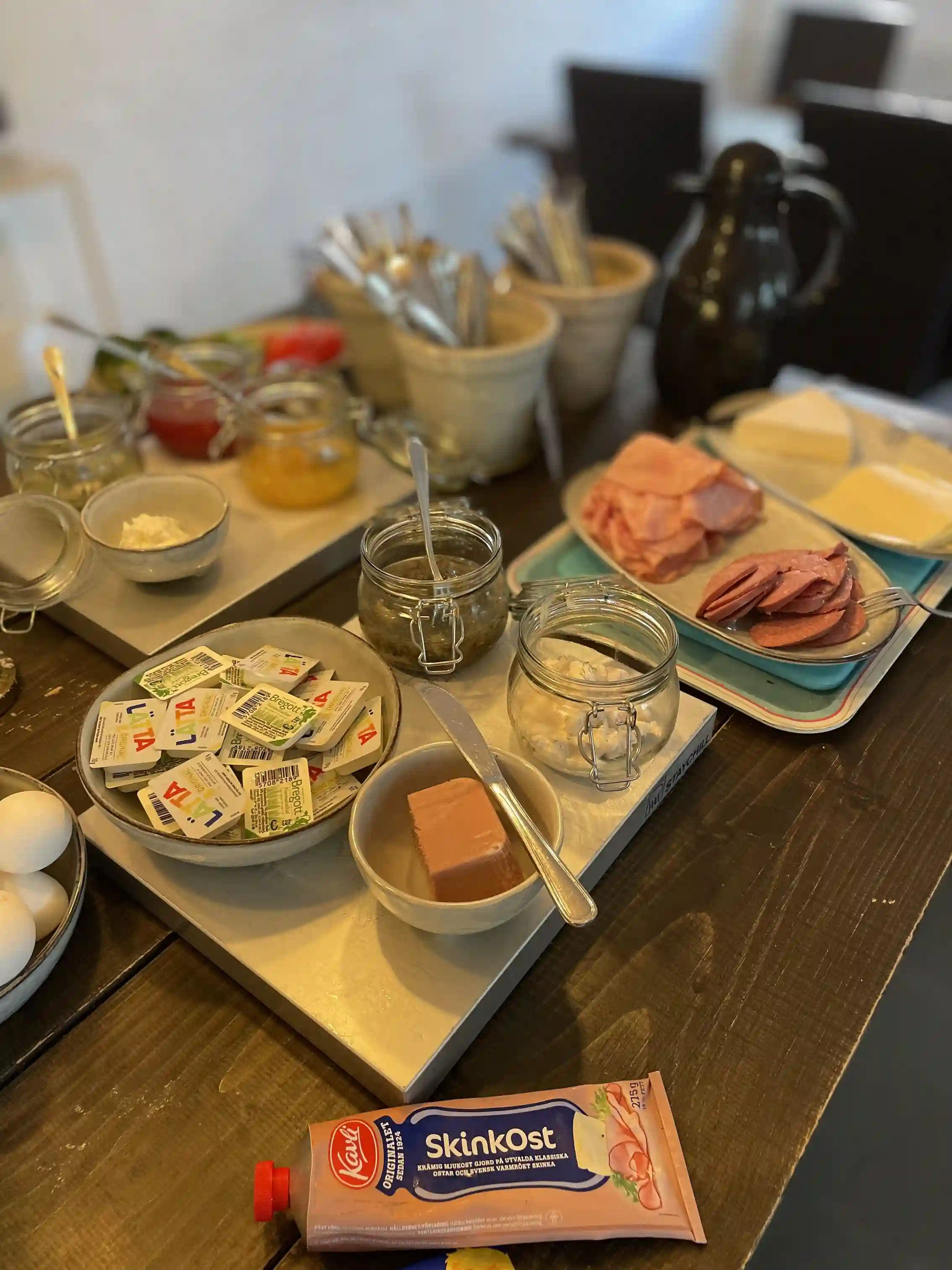 Bild på ett frukostuppdukat bord med olika rätter och tillbehör, inklusive tallrikar med smörgåsar, en skål med yoghurt samt diverse pålägg och mejeriprodukter. Perfekt för en avkopplande start på dagen på Husargården.