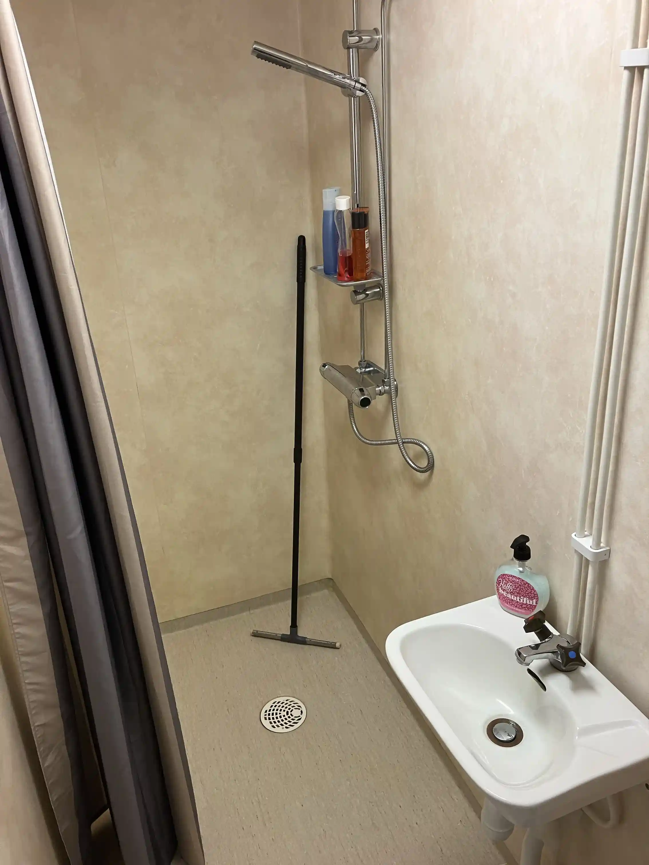 Bilden visar ett modernt badrum med kaklade väggar och golv. I centrum finns en dusch med en duschstång och ett regnduschmunstycke. Rör och annan badrumsarmatur framträder tydligt.