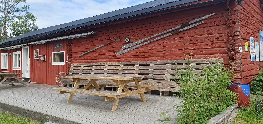 En stuga i trä ligger i en grönskande trädgård vid Kungsgårdsviken. Framför stugan står ett picknickbord av trä, omgivet av utemöbler, vilket skapar en inbjudande plats för avkoppling i naturen.