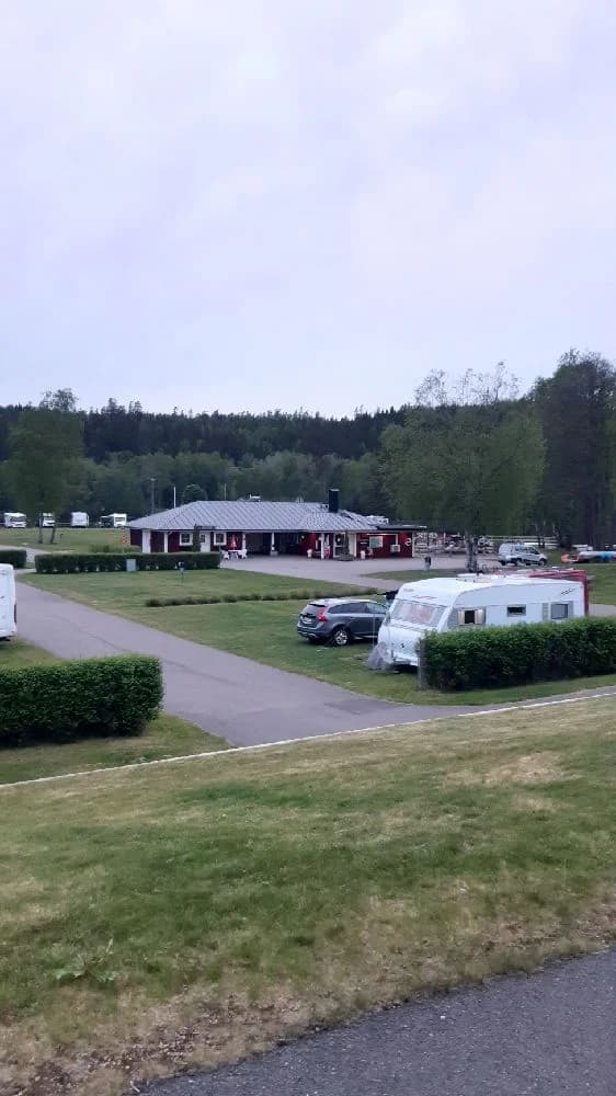 Bild av en parkeringsplats på Överlida camping med flera husbilar, husvagnar och bilar. Gräsmattor omger fordonen och ett skogsområde syns i bakgrunden.