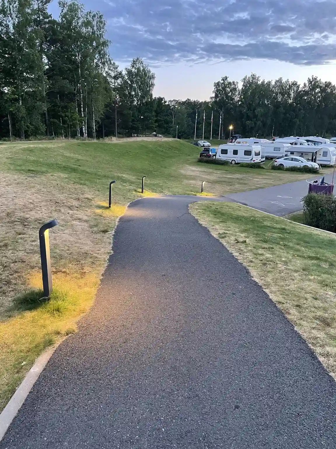 En asfalterad väg leder genom en lummig parkområde med grönskande gräsmattor och anlagda gångstigar vid Överlida Camping.