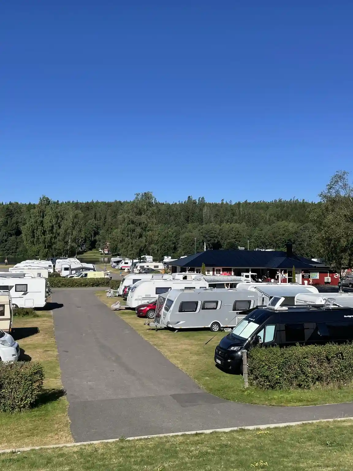 En bild över en campingplats med flera parkerade husbilar och husvagnar, omgivna av personbilar och ett mindre grönområde.