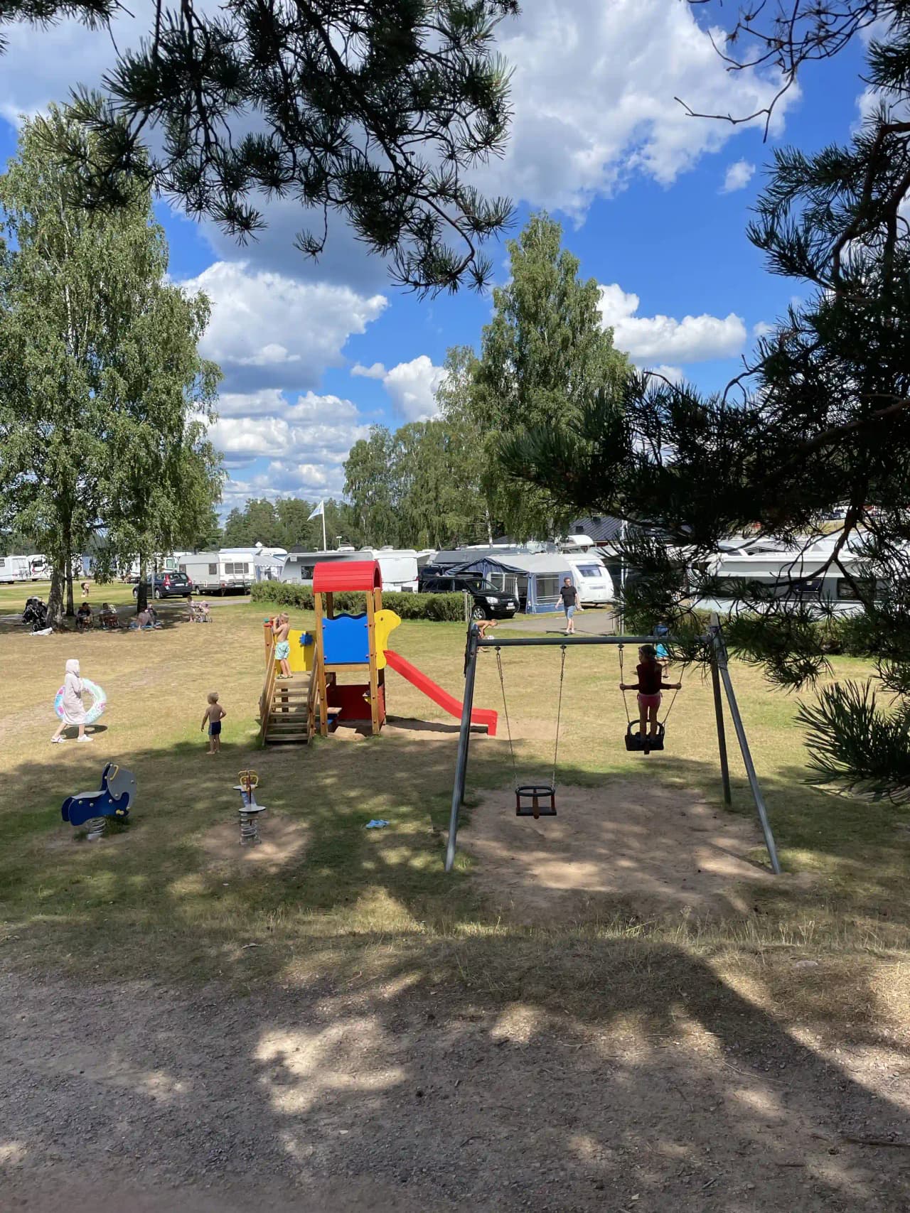 En bild av Overlida Camping med husbilar parkerade under skuggiga träd. I förgrunden syns en grön gräsmatta och en stolpe, vilket skapar en inbjudande atmosfär för rekreation och avkoppling i naturen.