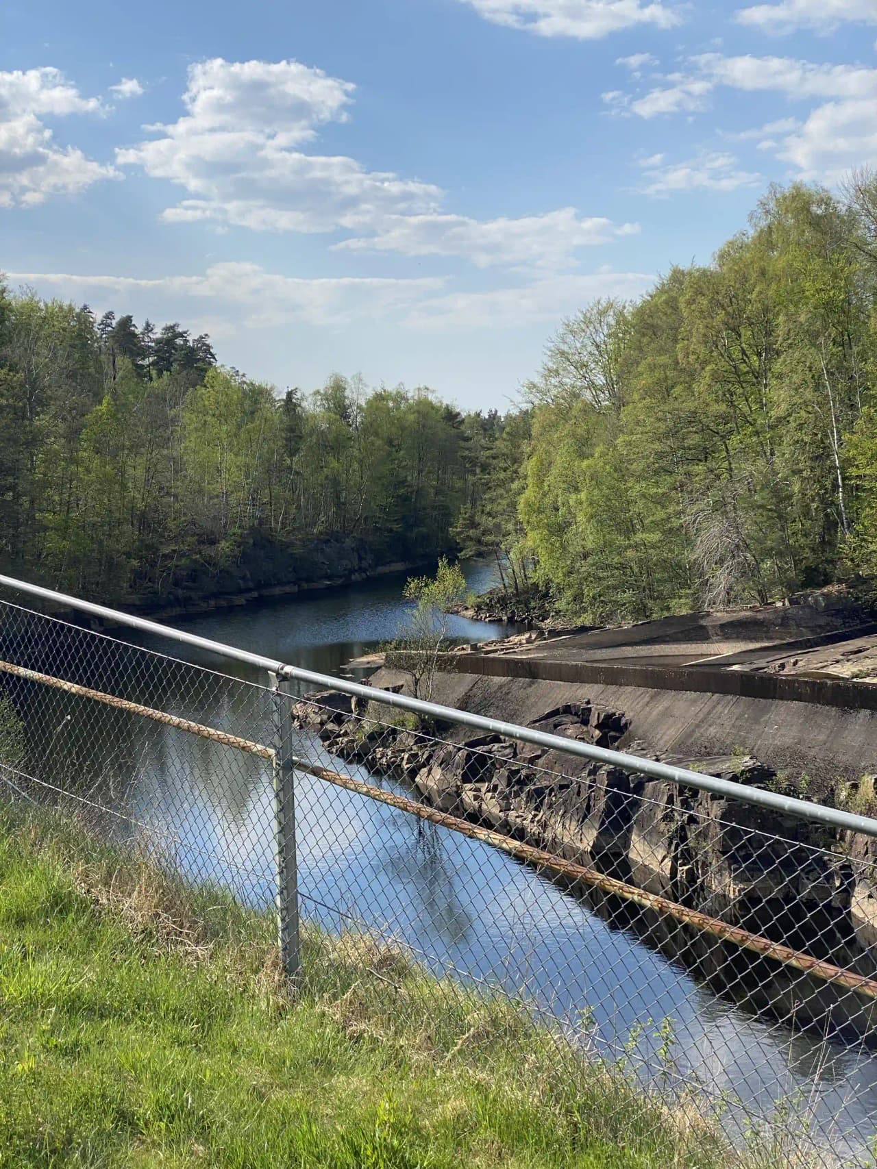 Bild på den Sovande Älgen: En vy av en metallnät-bro över en flod- eller kanalformation, omgiven av kedjelänkstängsel med grönska i bakgrunden.
