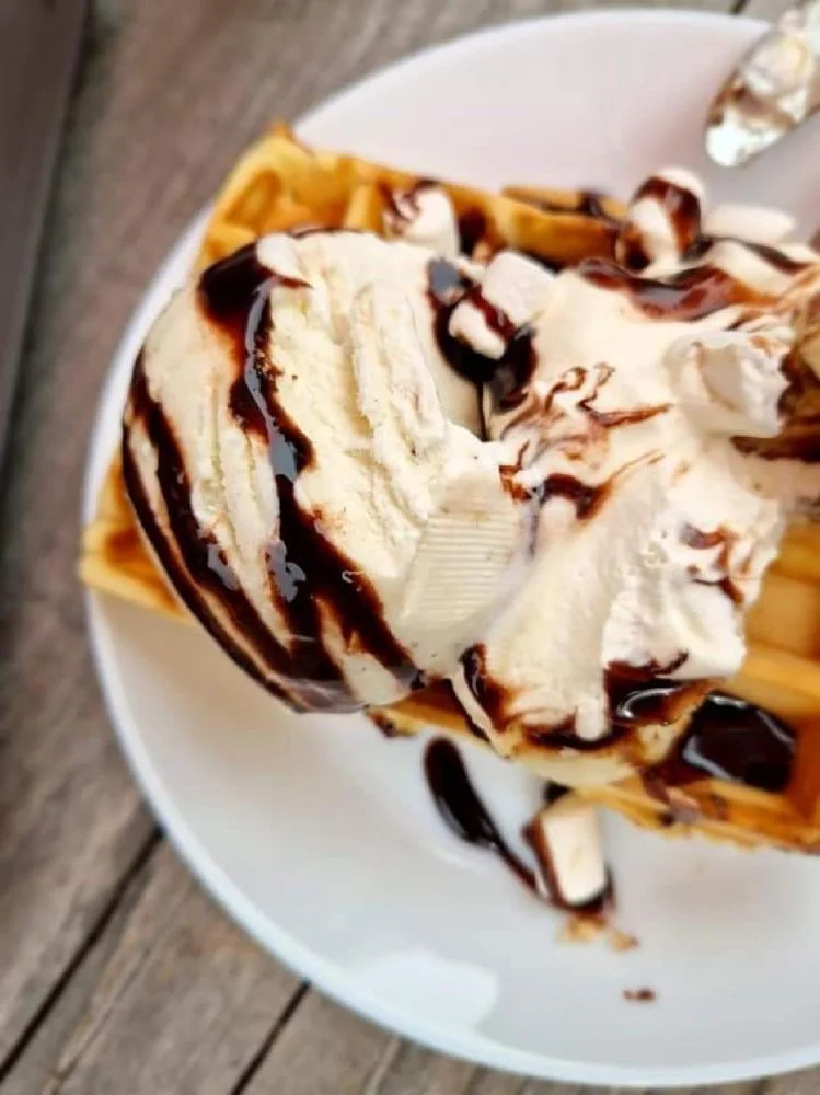En närbild av en belgisk våffla toppad med glass och chokladsås, omgiven av olika matingredienser och desserter, serverad på Hamra Stugby.