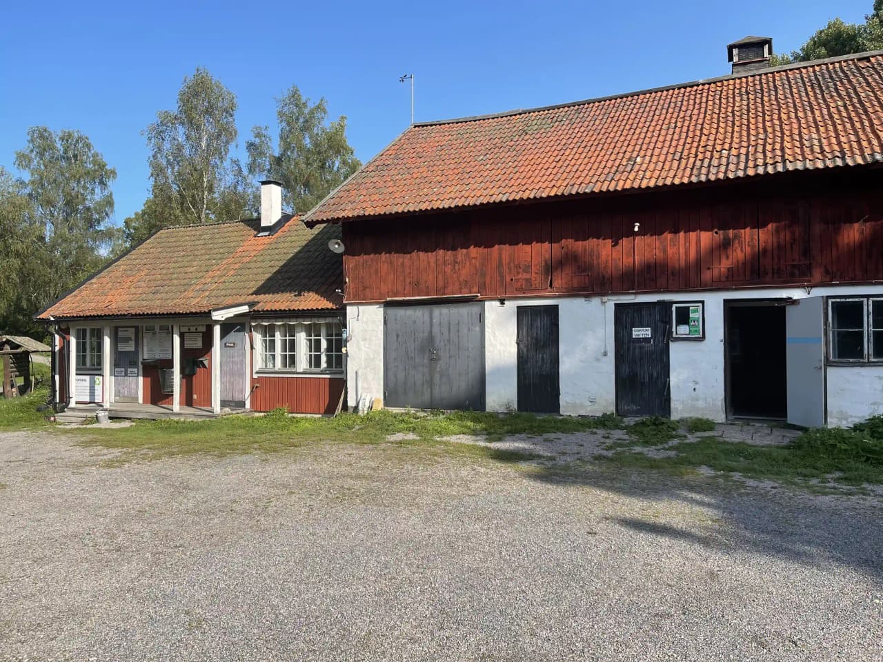 Bild av en lantlig miljö i Fyrväpplingen med ett litet hus med yttertak och panel, omgivet av flera små byggnader som en lada och skjul. En grusväg leder genom byn, omgiven av grönska och en fridfull landsbygd.
