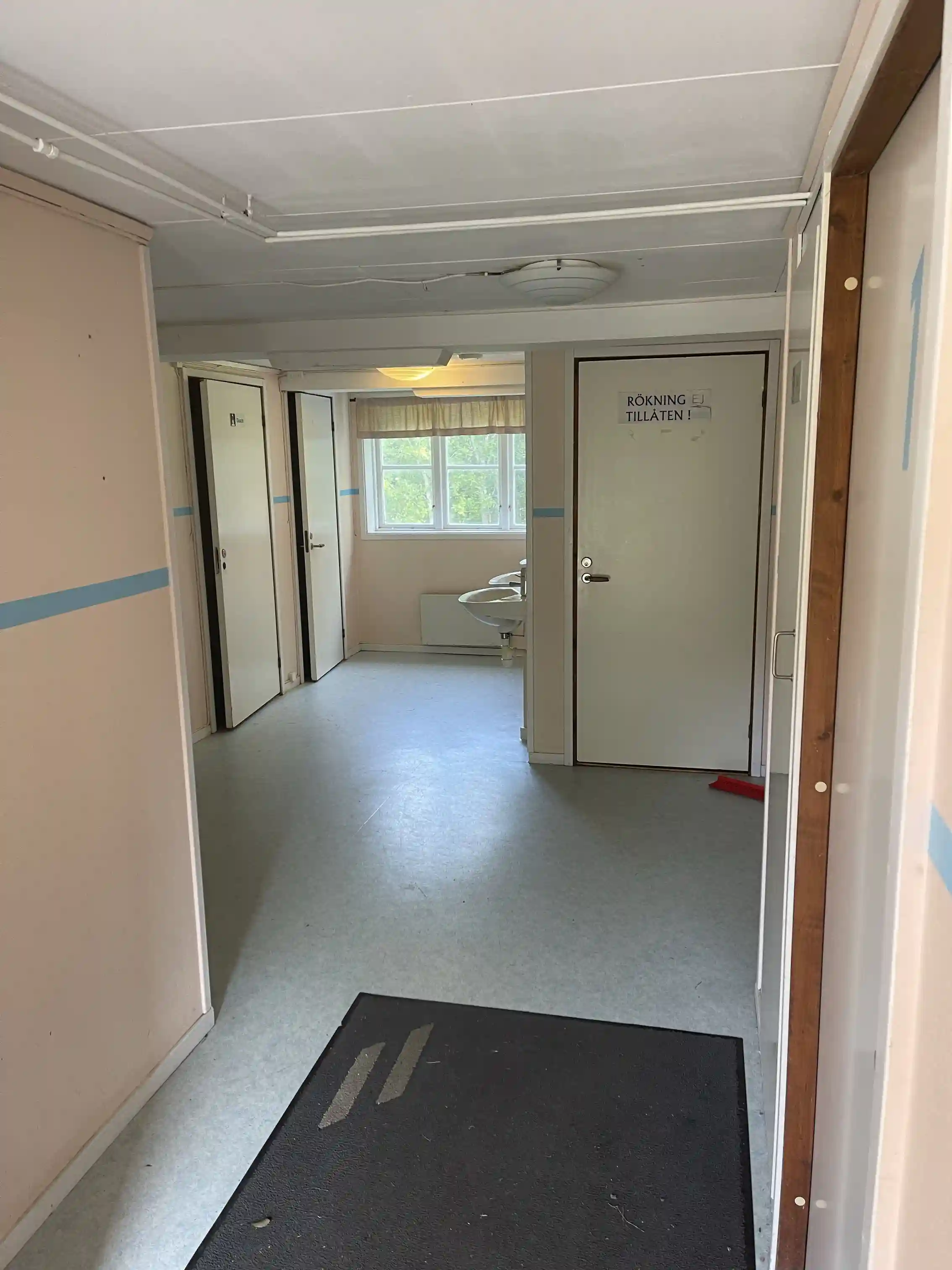 Ett ljust och modernt rum med kakelgolv, vita väggar och ett tak med infälld belysning. En dörr i aluminium leder ut från rummet och dagsljus strömmar in. Rummet ger ett intryck av renlighet och stilren design.