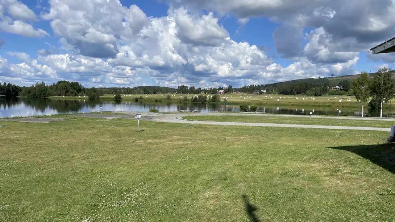 En somrig naturbild som visar en vattenmassa med omgivande strand och grönska, ackompanjerad av en klar himmel med mjuka cumulusmoln.