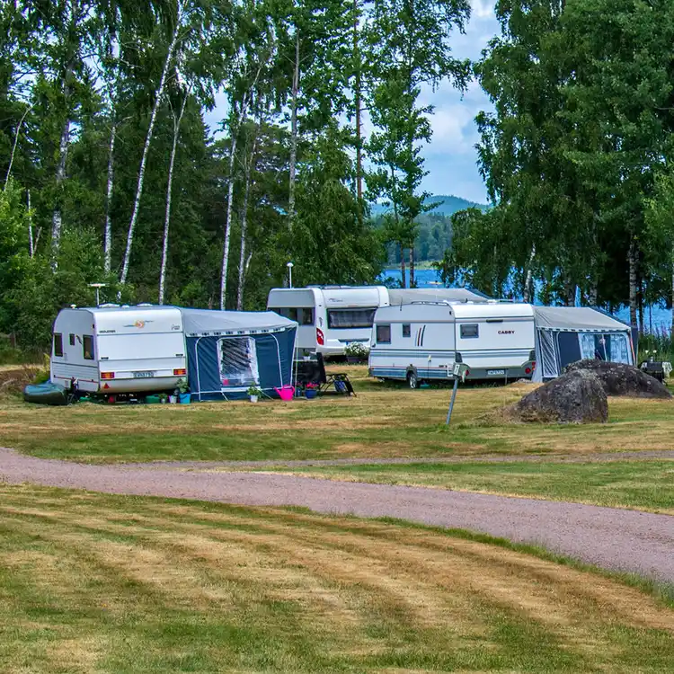 Vy över Lungsunds camping med grönskande träd och gräs. Flera husvagnar och tält är uppställda på en plan yta omgiven av öppna fält och natur.