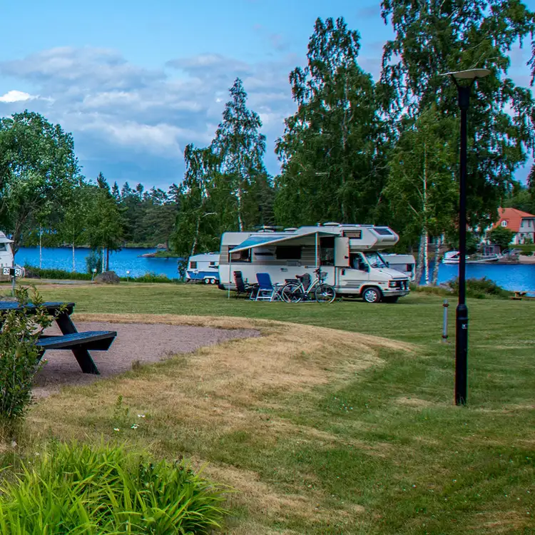 En campingplats vid en sjö med husvagnar och en skåpbil parkerade på gräset. I bakgrunden syns träd och en klarblå himmel.