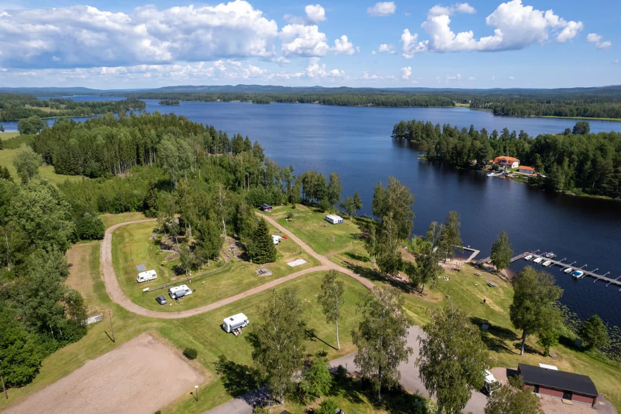Flygfoto över Lungsunds camping med stugor vid sjöns strand. Omgivande landskap inkluderar skog och flod i en naturskön miljö.