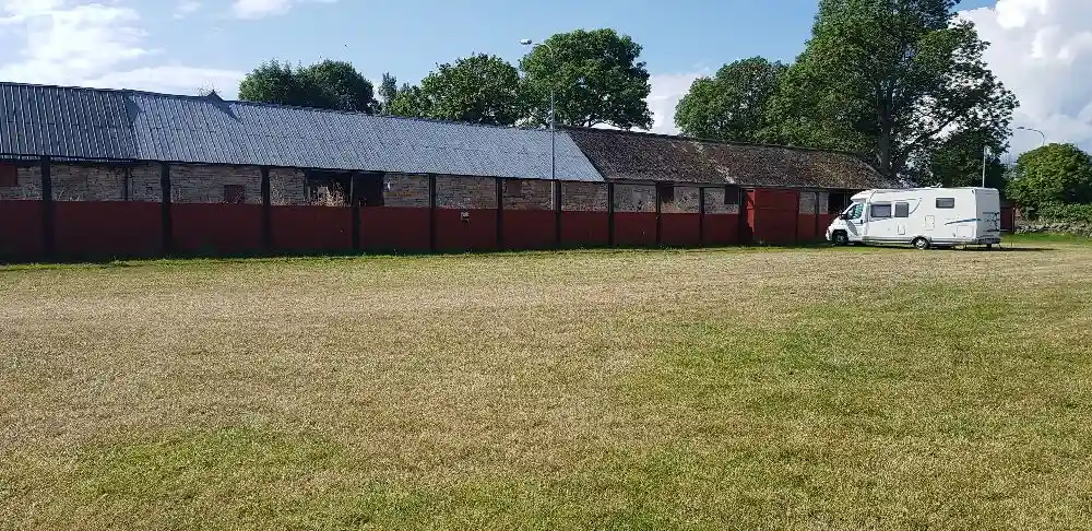 En grön äng i ett lantligt område vid Kalvhagen, Södvik, med gräs och betesmark i förgrunden. I bakgrunden syns några byggnader med tak som ligger utspridda bland träden.
