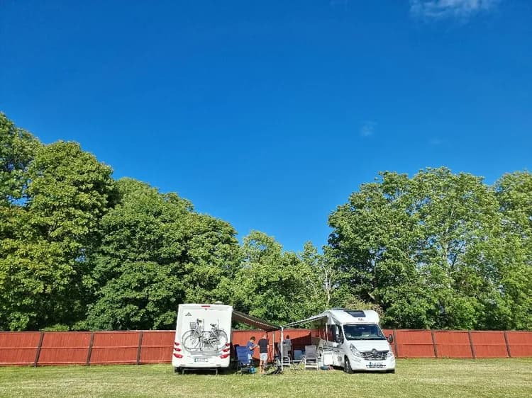 En grön gräsmatta med flera tält uppställda i ett campingområde. En husbil och en skåpbil står parkerade intill, omgivna av träd.