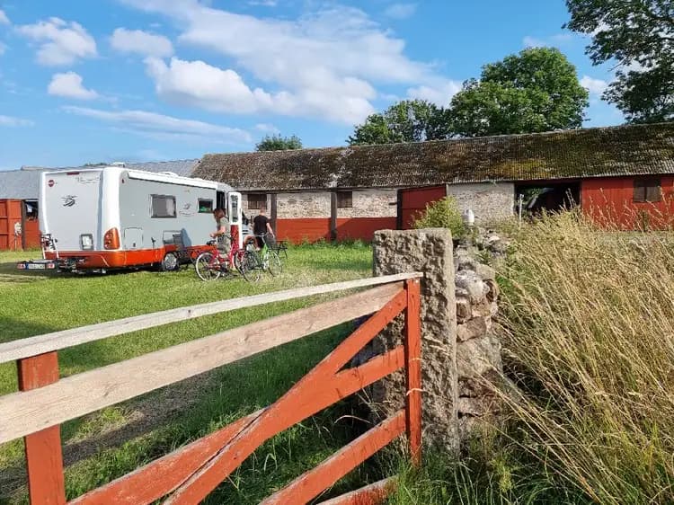 En lantlig miljö i Kalvhagen, Södvik, med ett rött torp omgivet av vidsträckta gräsytor och lantlig bebyggelse. I bakgrunden syns ett vitt uthus och flera träd, vilket förstärker den idylliska och genuina landskapskänslan.