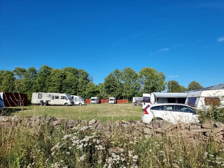 En campingplats i Södvik, Kalvhagen, med flera husbilar och husvagnar parkerade i en stor grusbelagd parkeringsyta omgiven av gröna träd och buskar.