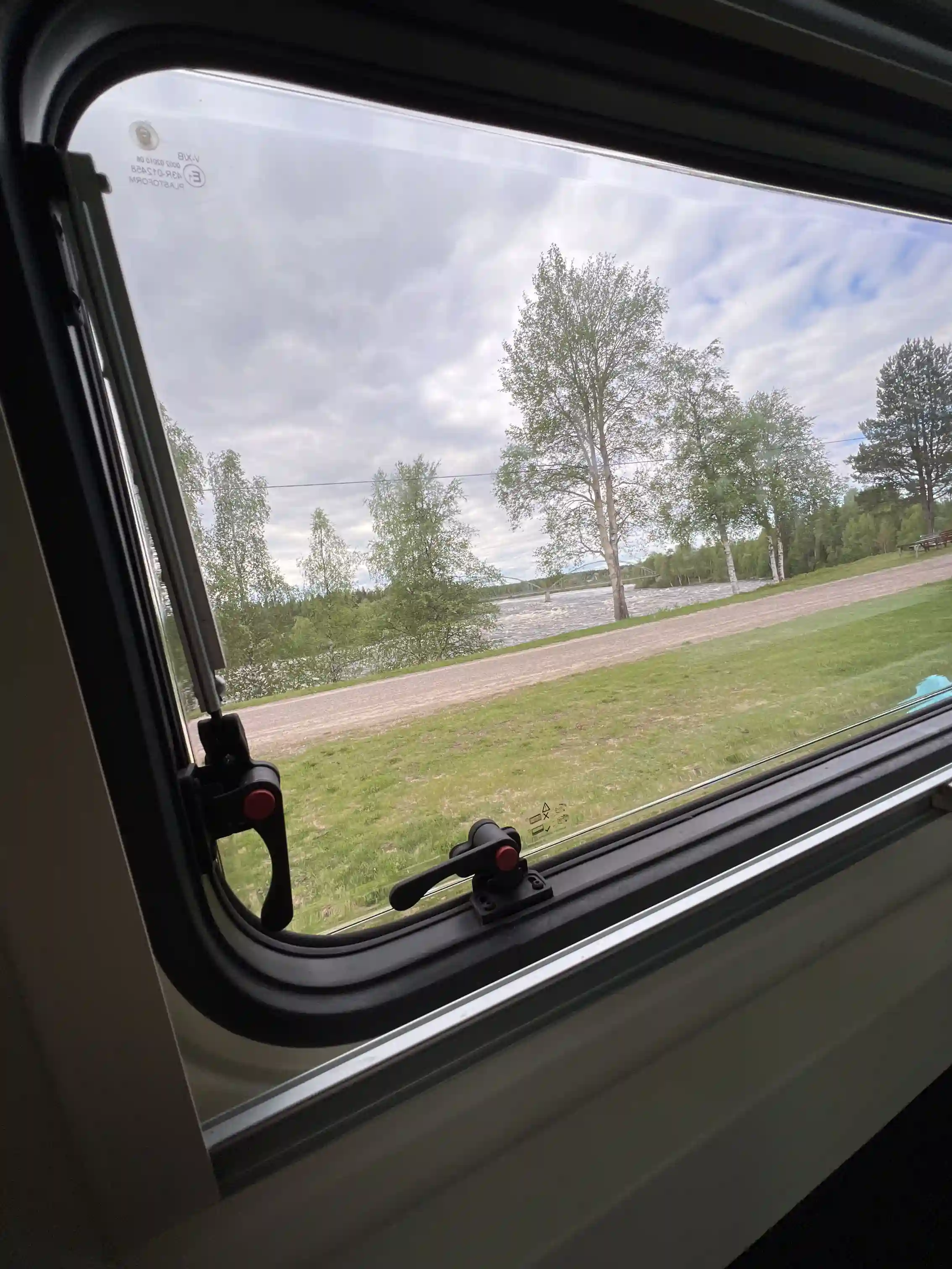 En öppet landskap med gräsfält och en väg leder mot Kamlunge Camping, där en bil med glass på vindrutan är synlig i förgrunden.