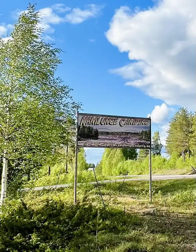 En naturskön vy över en campingplats vid Kamlunge, omgiven av frodig grönska och träd. En skylt visar vägen till platsen. Morgonsolen silar genom grenverket och skapar en lugn och fridfull atmosfär.