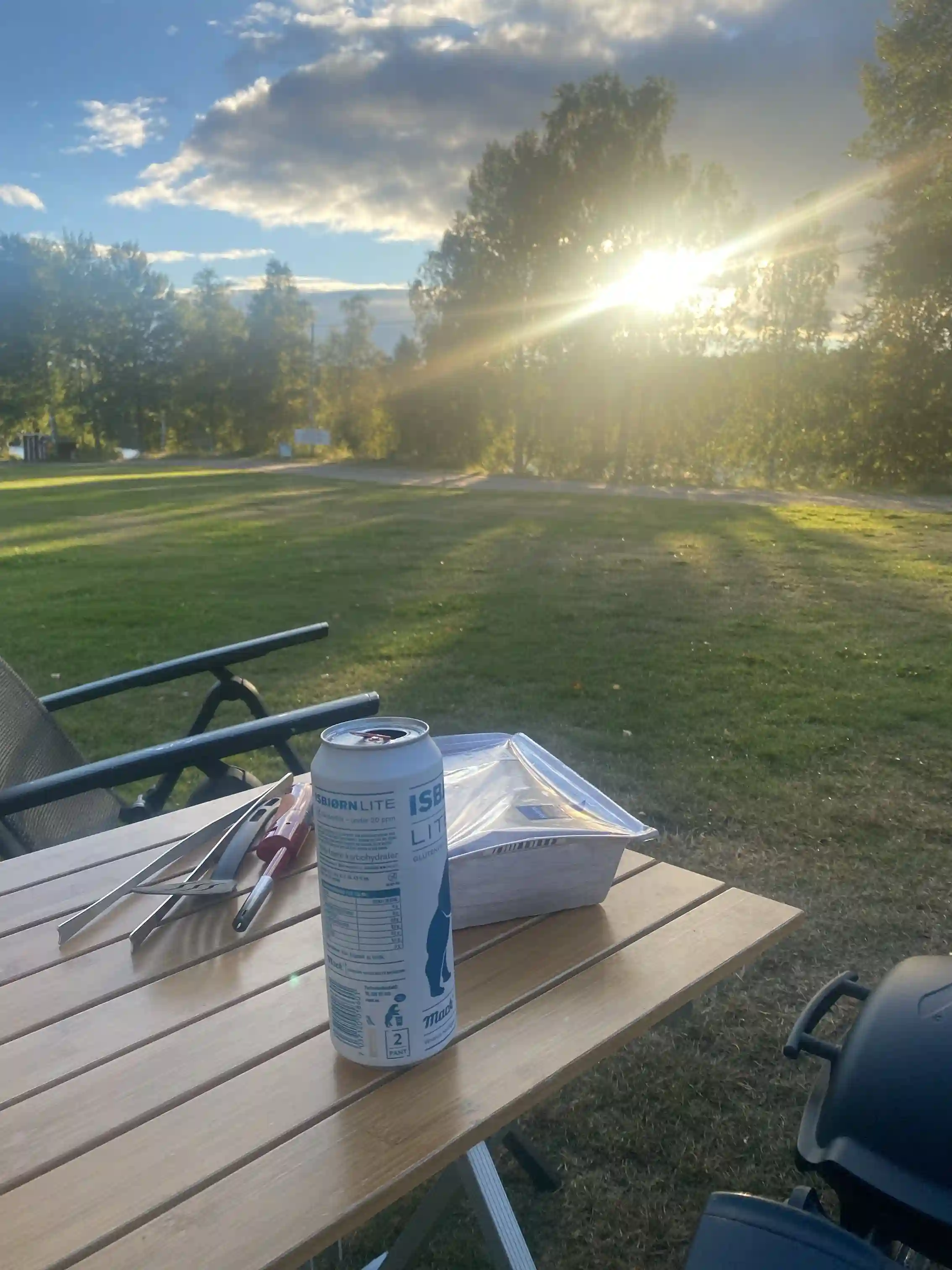 En utomhusutsikt över Kamlunge Camping med trämöbler under skugga. Solbelysta picknickbord och stolar ger en inbjudande atmosfär för besökare.