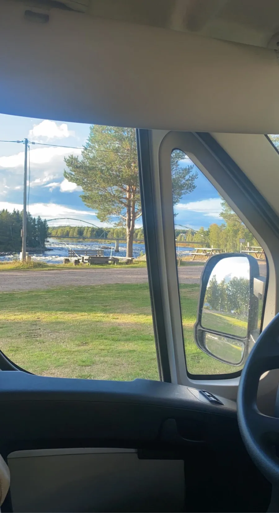 En solig morgon vid Kamlunge camping, sedd genom en bilruta. Solens strålar lyser över landskapet, med bilens speglar och dörr synliga i förgrunden, vilket skapar en känsla av ett pågående roadtripäventyr.