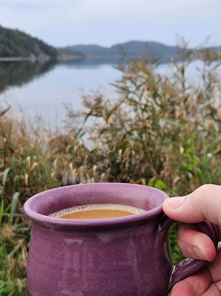 En vit kaffekopp fylld med rykande kaffe står på ett dukat bord vid Vindöns Camping och Marina. I bakgrunden syns en naturskön miljö som ger en avkopplande atmosfär till platsen.