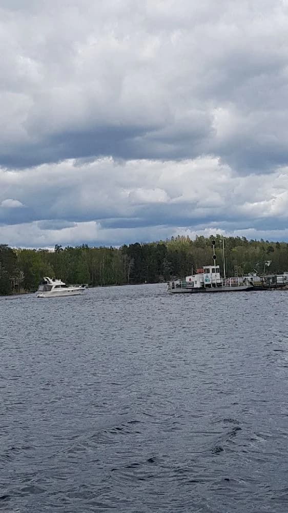 En naturskön vy över en kanal med flera småbåtar på vattnet, omgivna av grönskande landskap och en molnig himmel, på Hätte Camping.