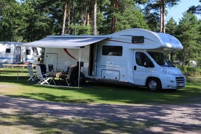 En husbil står parkerad vid en campingplats, omgiven av grönska. I förgrunden syns en bil med öppen dörr, och i bakgrunden skymtar ytterligare en husbil samt en campingvagn. Det är soligt och området ger en känsla av avkoppling och utomhusliv.