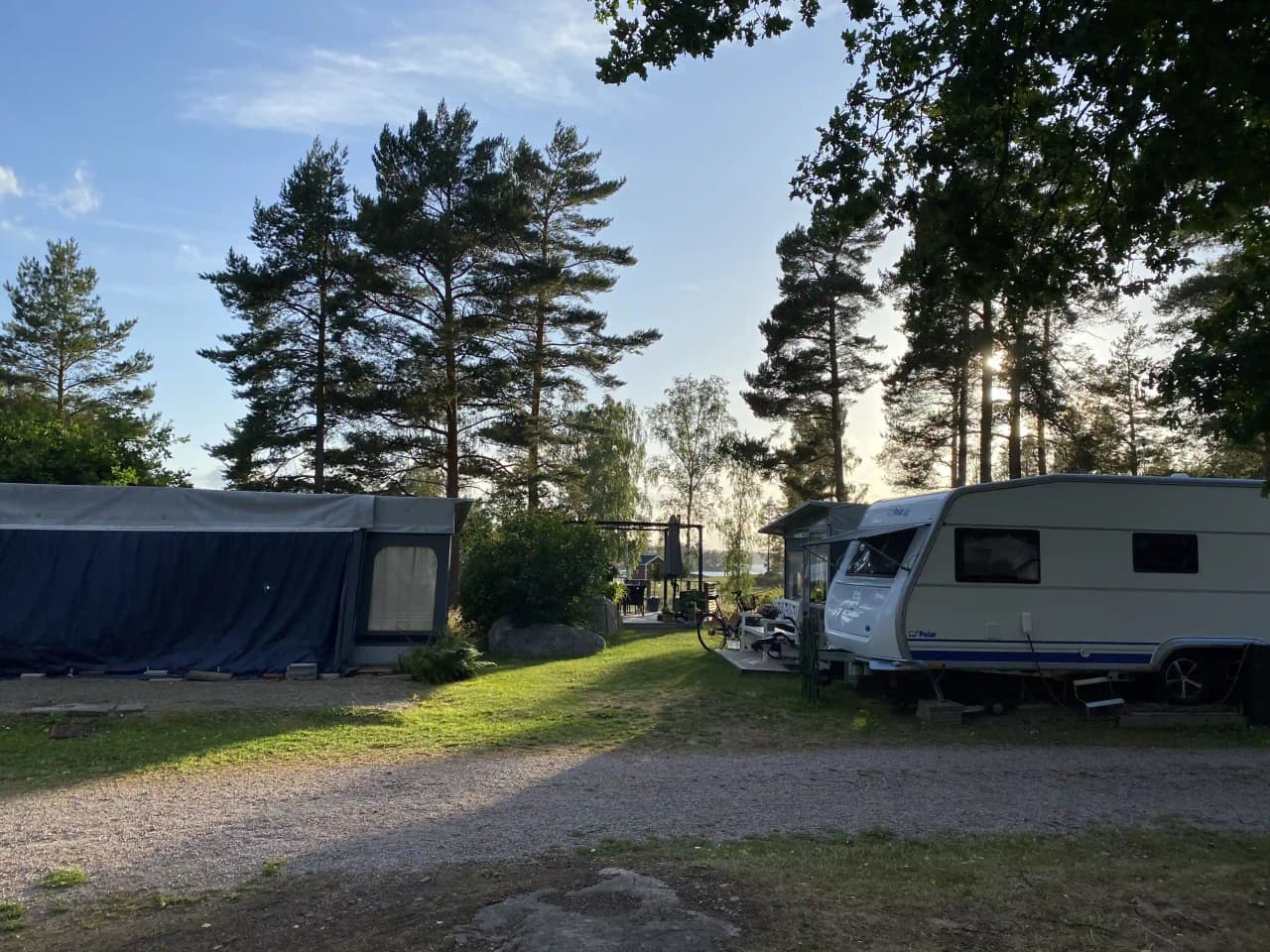 En campingplats med husvagnar och husbilar uppställda bland träd och växter, omgiven av en gräsbevuxen gård och parkerade fordon.