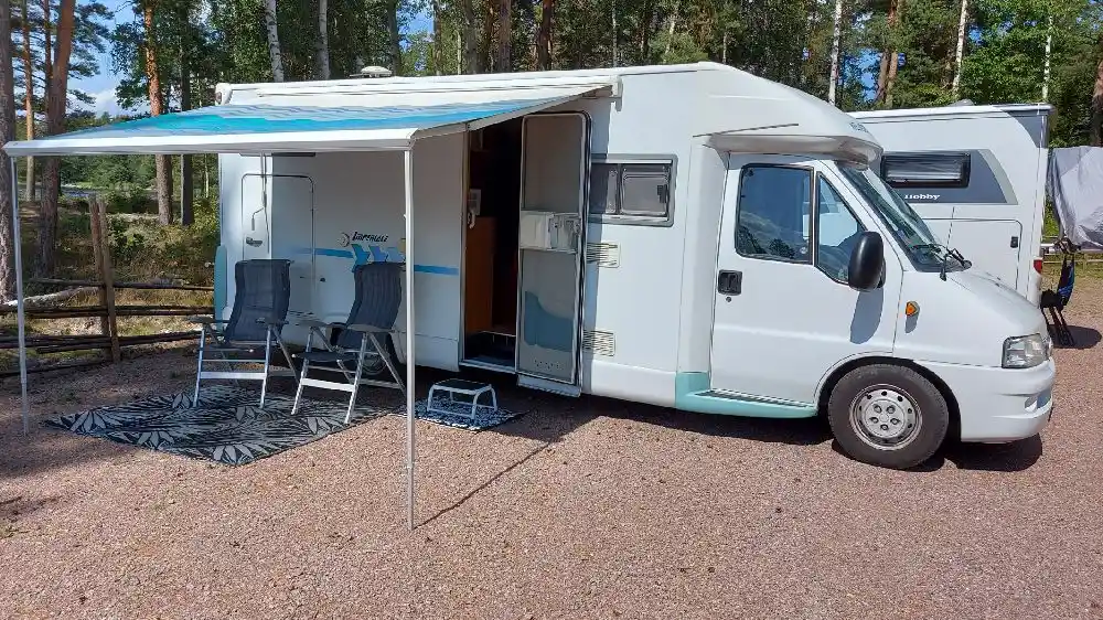En rad av husbilar och husvagnar uppställda på en campingplats, omgivna av grönska och grusvägar.